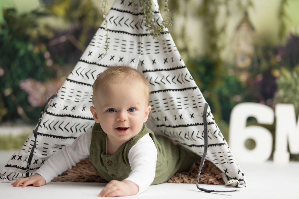 Smiling baby at 6 months old poses in front of a white teepee tent during a milestone photo session.