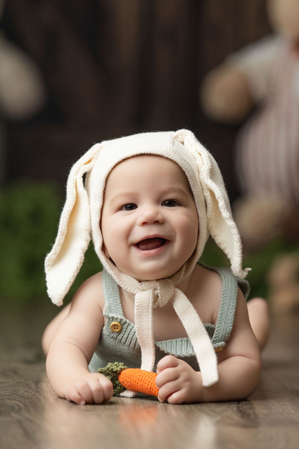 Smiling baby in white bunny ears hat and green overalls holding a crochet carrot toy on wooden floor.