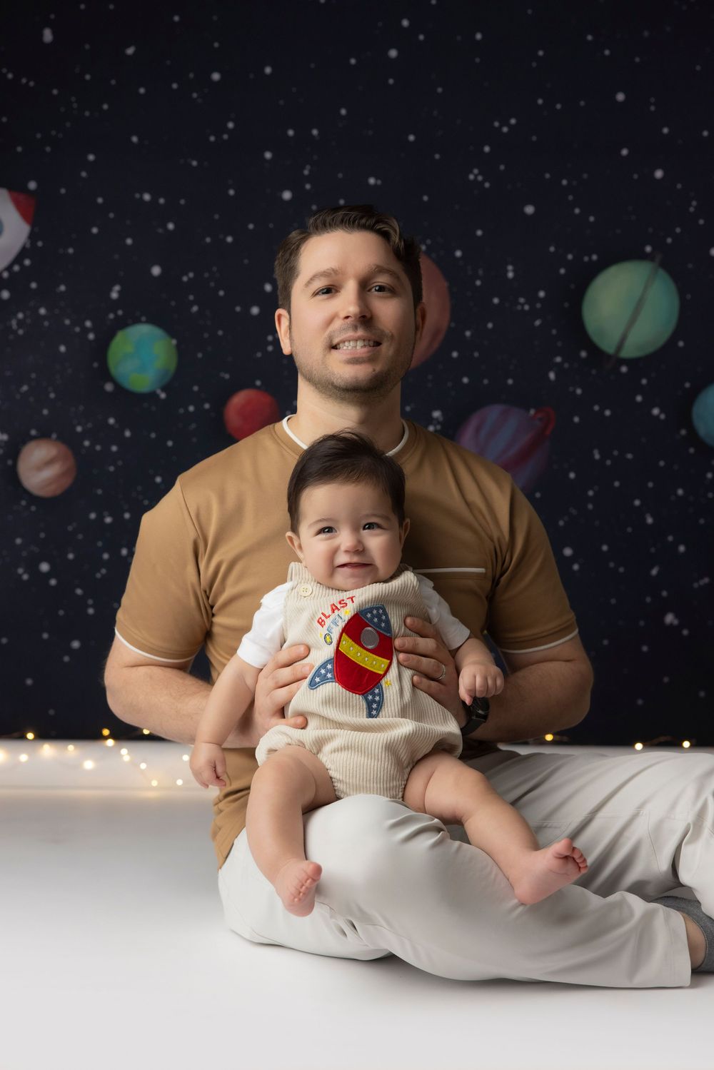 Father sitting cross-legged holding smiling baby in rocket outfit against space-themed backdrop with planets.