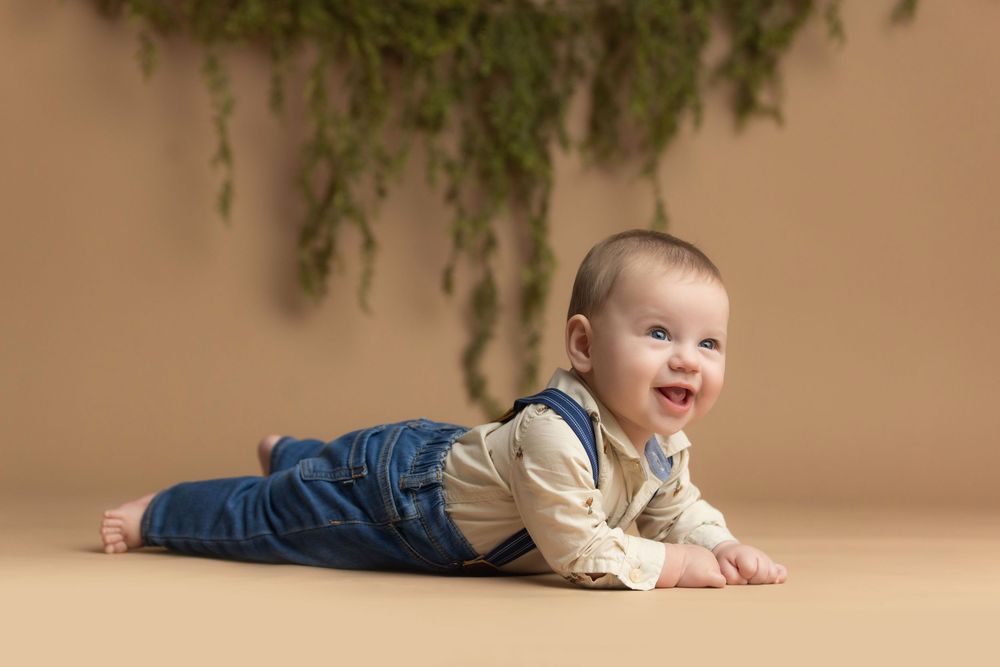 Smiling baby in denim overalls lying on stomach on beige floor with green hanging plants in background.