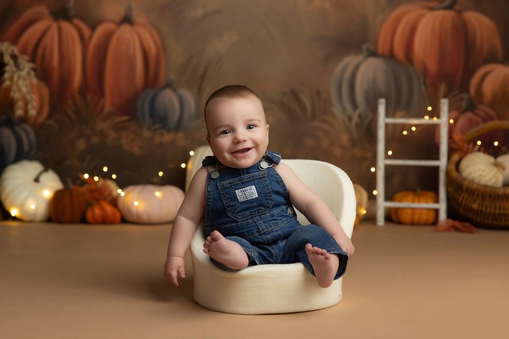 Smiling baby in denim overalls sits in a cream chair with a fall pumpkin backdrop and warm fairy lights.