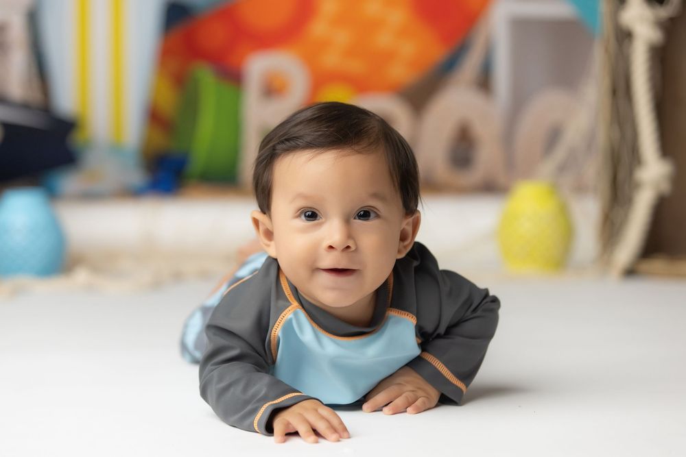 Cute baby boy in gray and blue outfit doing tummy time on white floor with colorful backdrop.