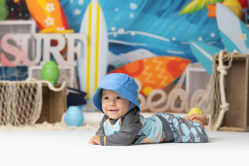 Adorable baby boy wearing blue hat doing tummy time in front of colorful surf-themed backdrop.