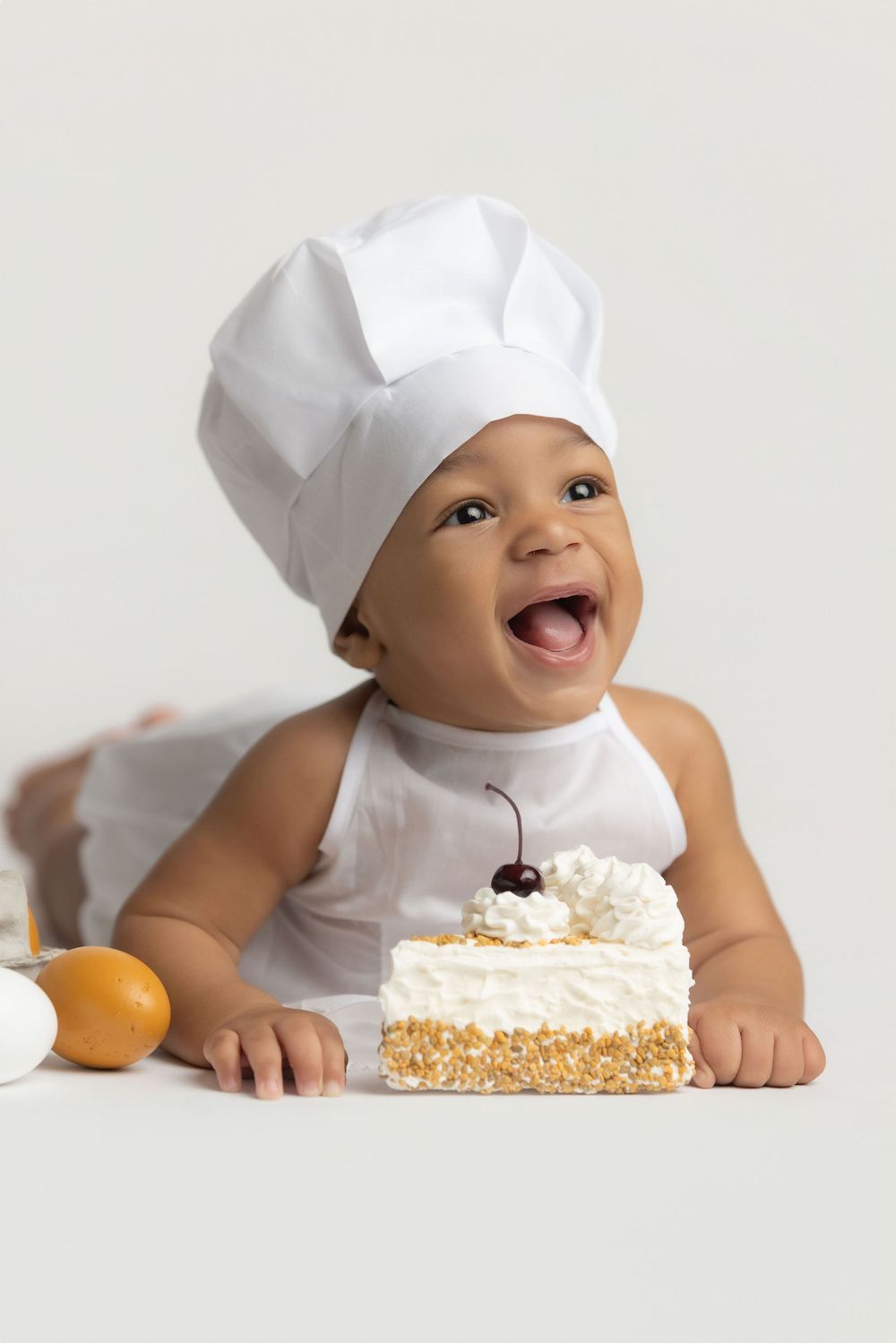 Adorable baby in a white chef hat smiling beside a decorated cake with cherry on top.