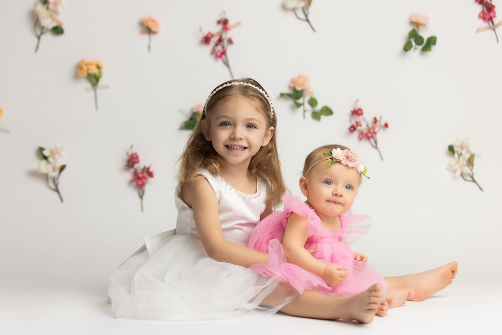 Two young sisters in white and pink dresses pose together against a floral wall backdrop in a studio portrait.