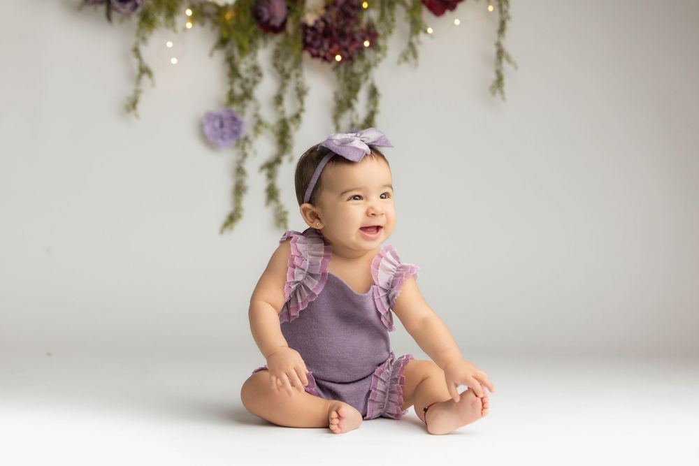 Smiling baby girl in purple romper and bow headband sits on white floor with floral backdrop.