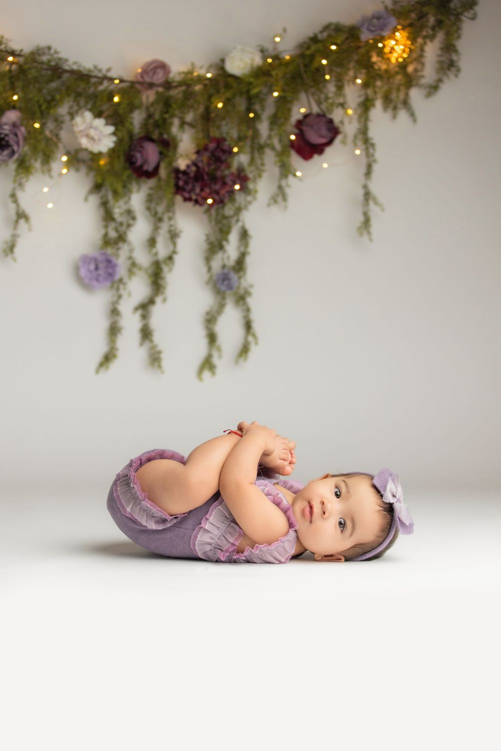 Baby girl in purple romper lying on white floor beneath floral garland with fairy lights.