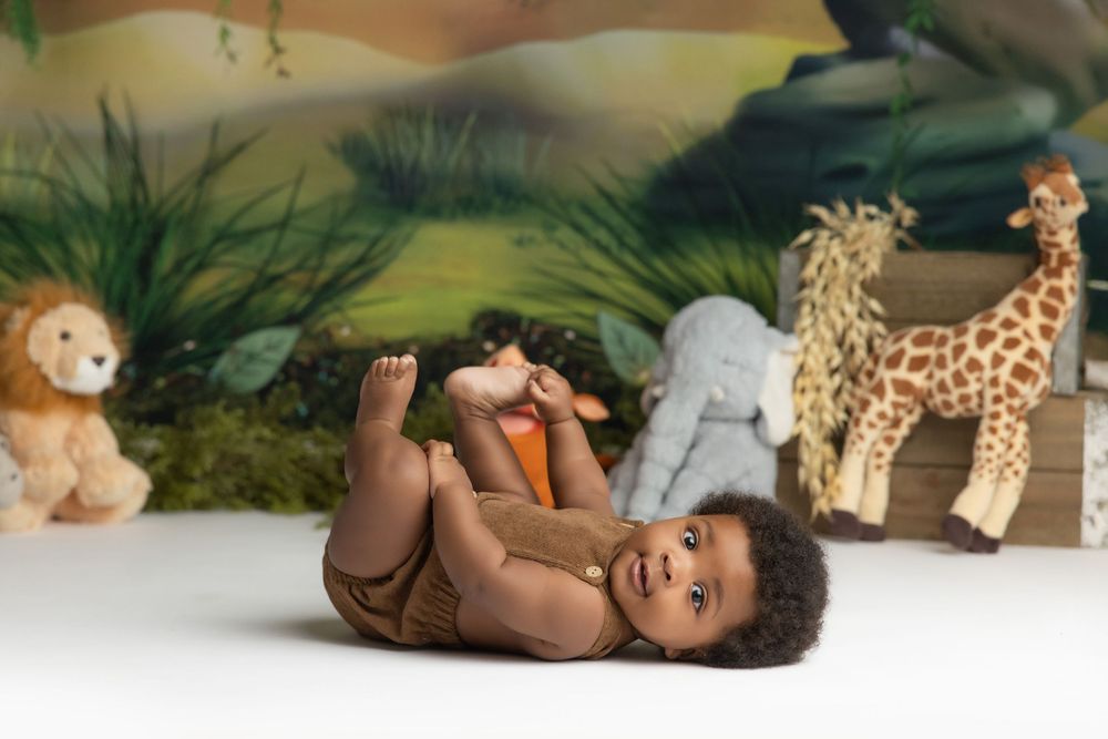 Smiling baby lying on back surrounded by stuffed safari animals including a giraffe and lion in a jungle-themed photo setting.
