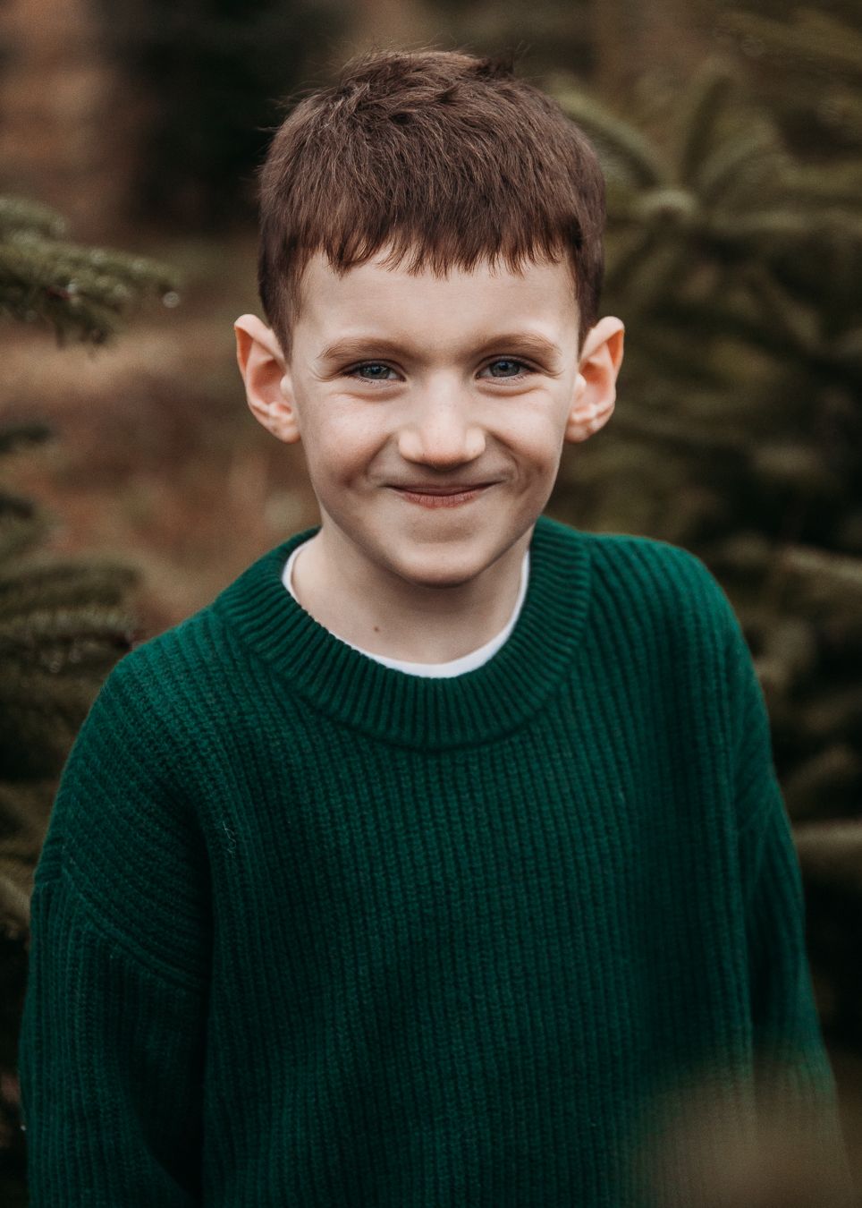 A young person in a dark green knit sweater smiles warmly against an outdoor background with evergreen trees.