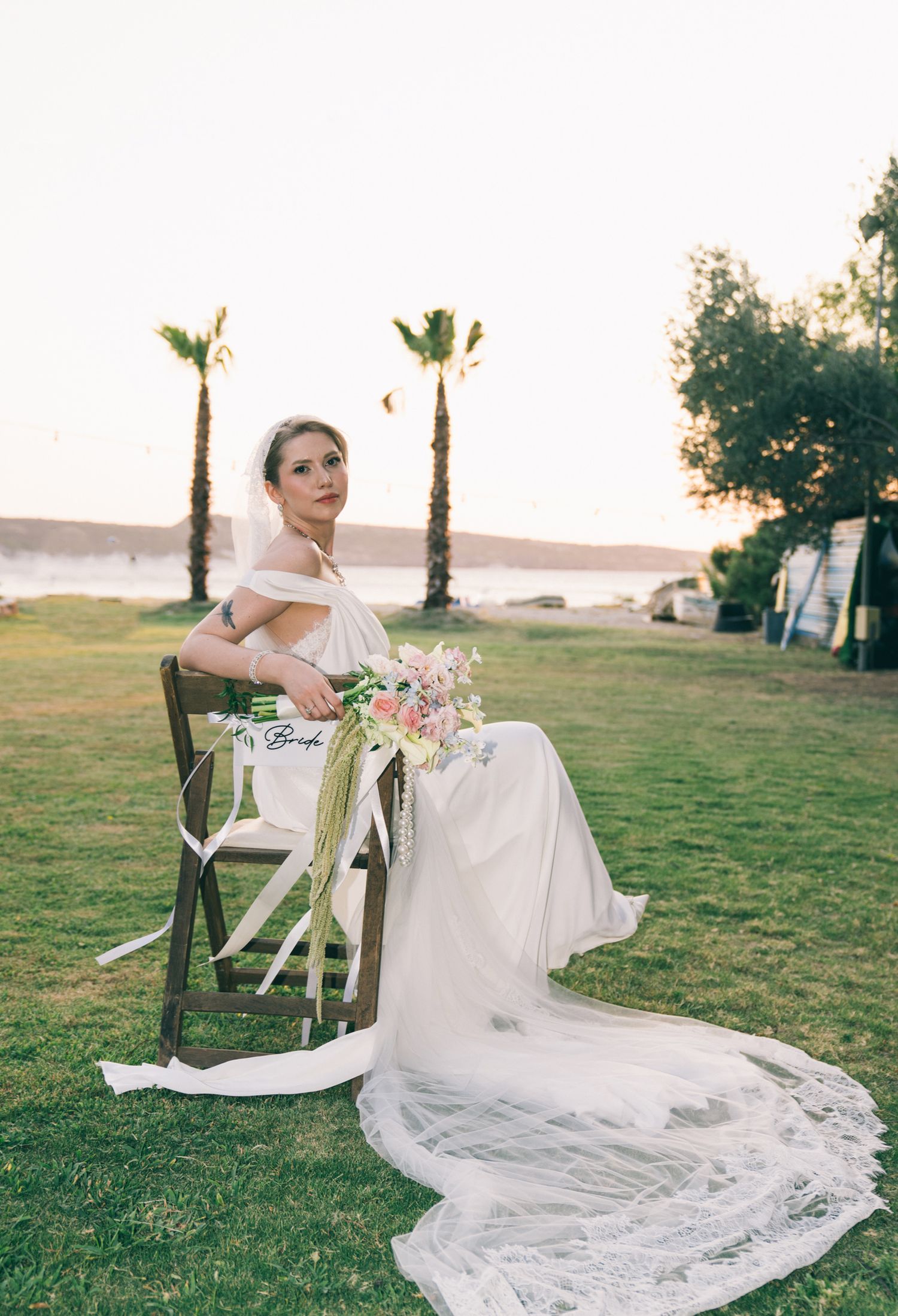 A bride in a flowing white dress sits on a chair on a lawn with palm trees and water views at sunset.