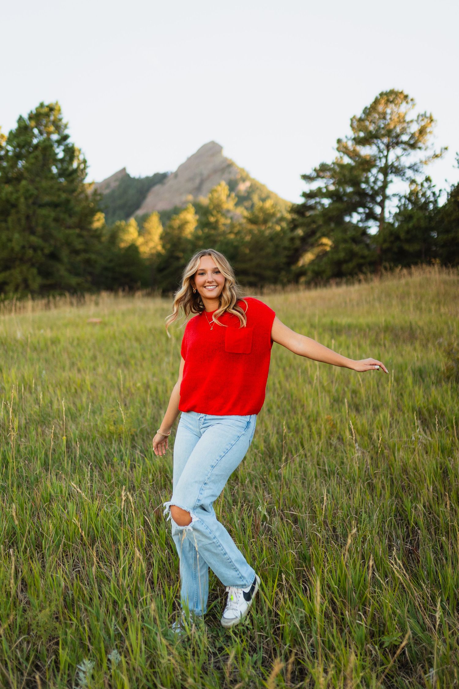 A person wearing a red top and ripped jeans dances joyfully through a sunlit meadow with mountains in the background.