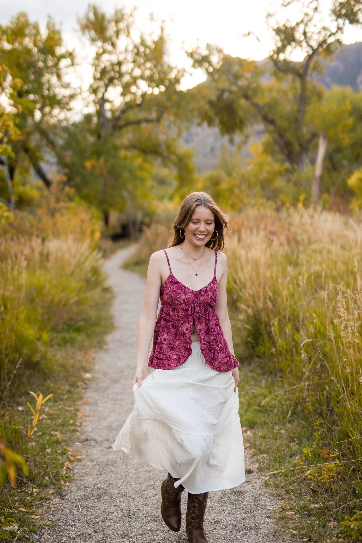 A person in a burgundy top and white skirt walks along a dirt path through tall grass and autumn foliage at sunset.