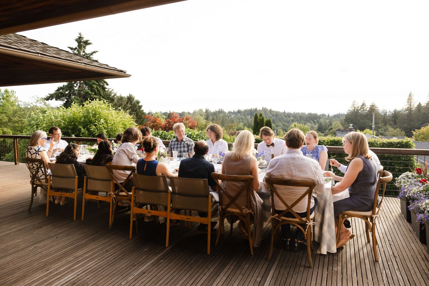 Wedding guests dine together at long wooden tables on an outdoor deck overlooking a scenic mountain landscape at sunset.