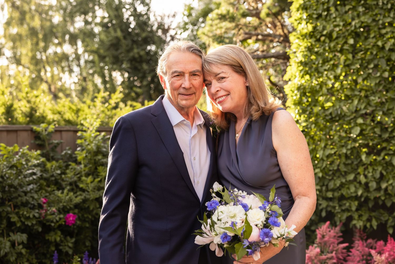 A couple stands together in a garden setting, with the woman holding a bouquet of purple and blue flowers.
