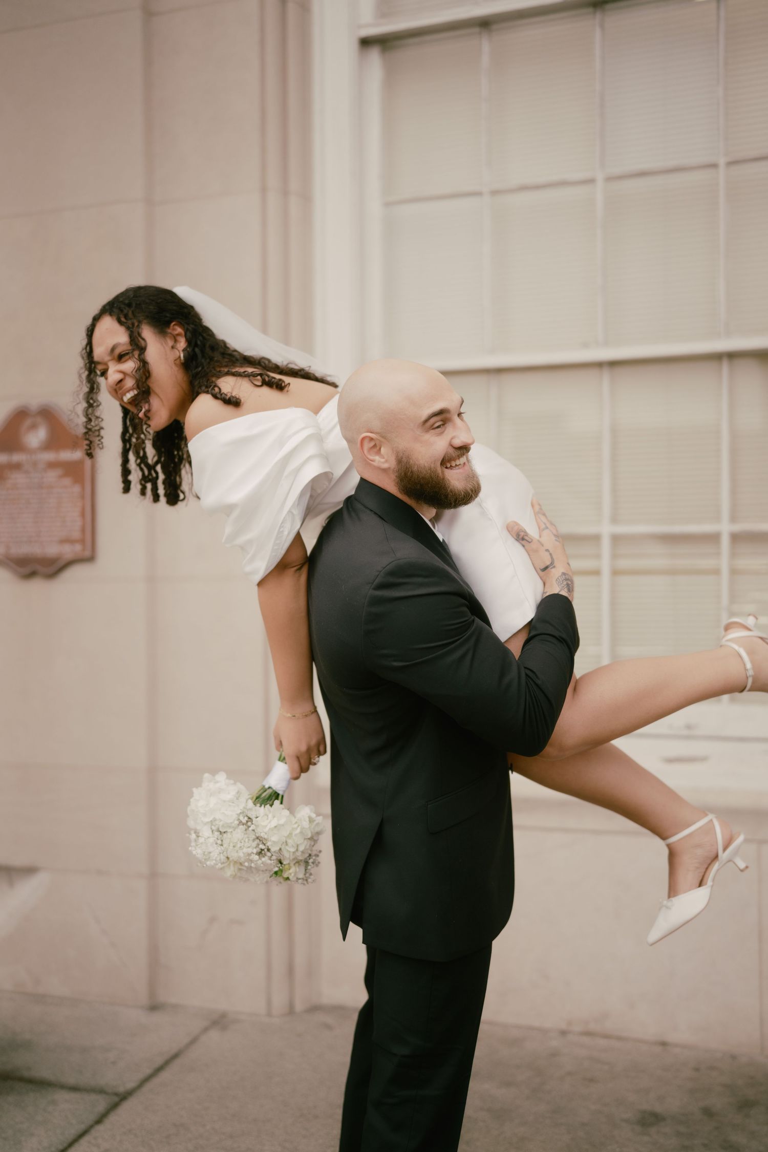 A joyful couple shares a playful moment outside as the groom carries the bride in her white dress and heels.