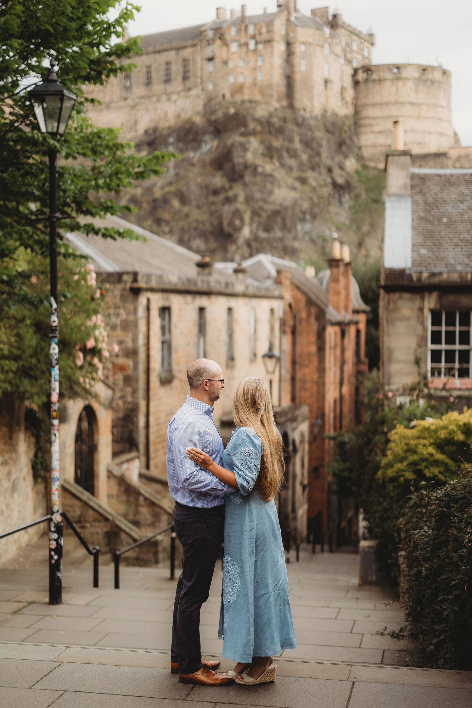 Two people walk together through a scenic stone courtyard with historic buildings and greenery in the background.