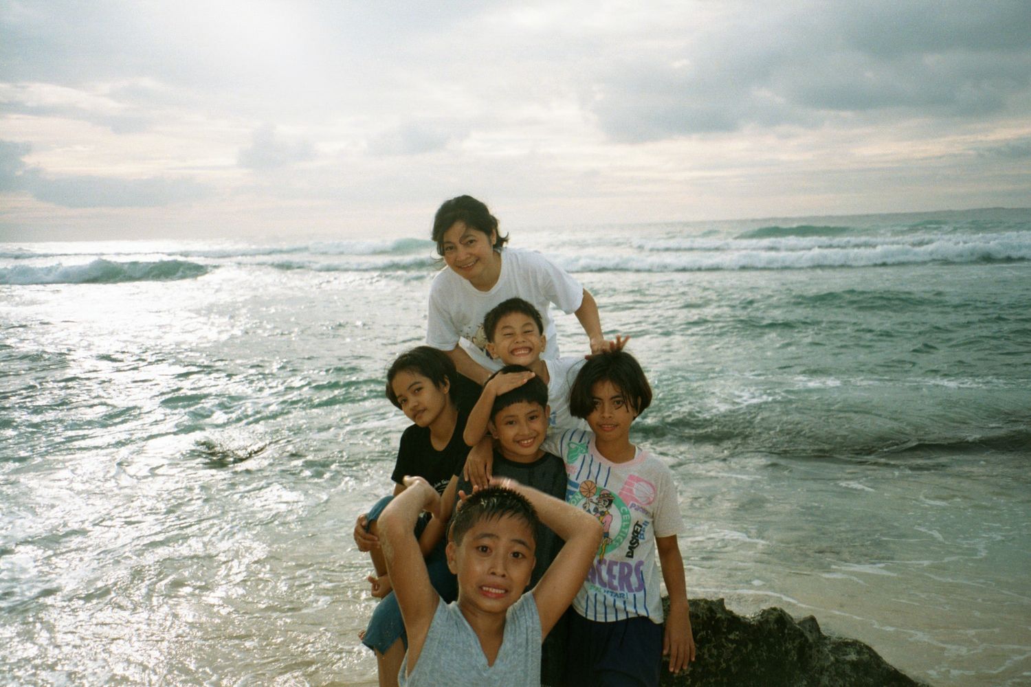 A group of people pose together joyfully at the beach with waves crashing behind them during sunset.