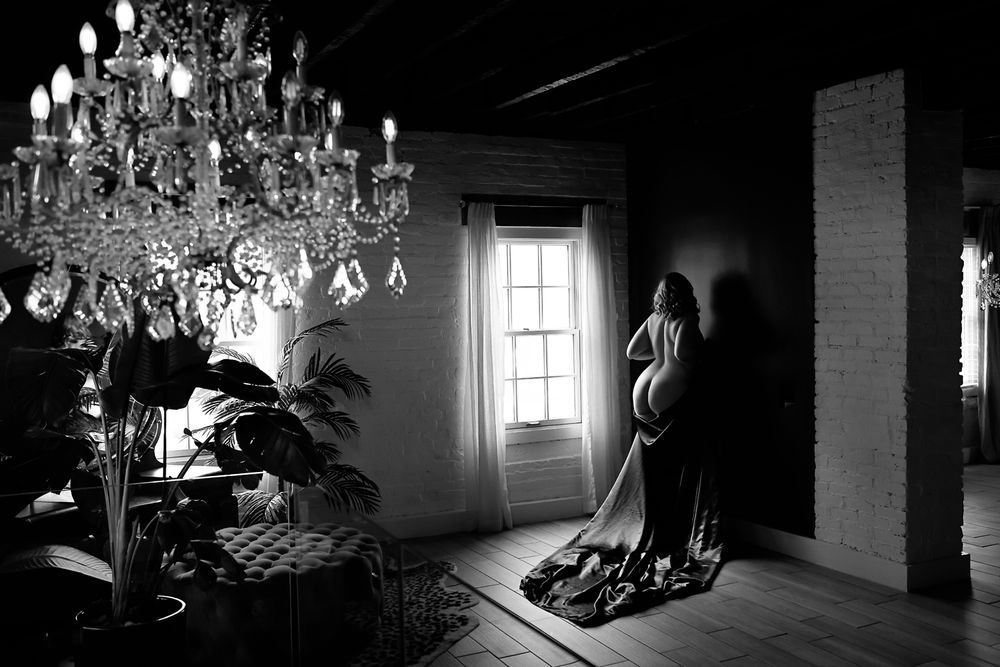 A dramatic black and white interior scene features an ornate crystal chandelier and figure standing by a sunlit window.