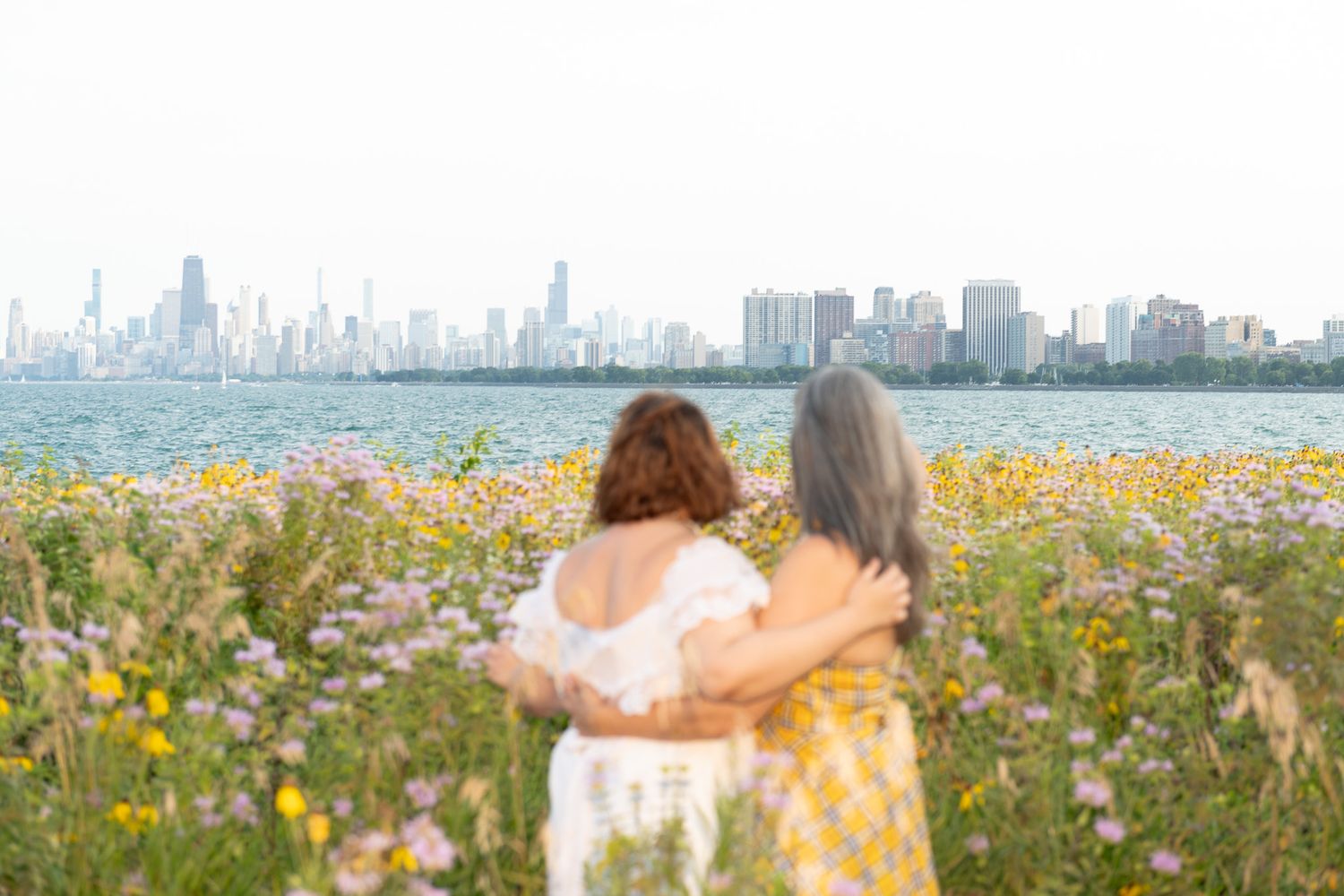 Two unknown women in dresses matching a floral field in bloom look over water to Chicago. A misty view of the city skyline.