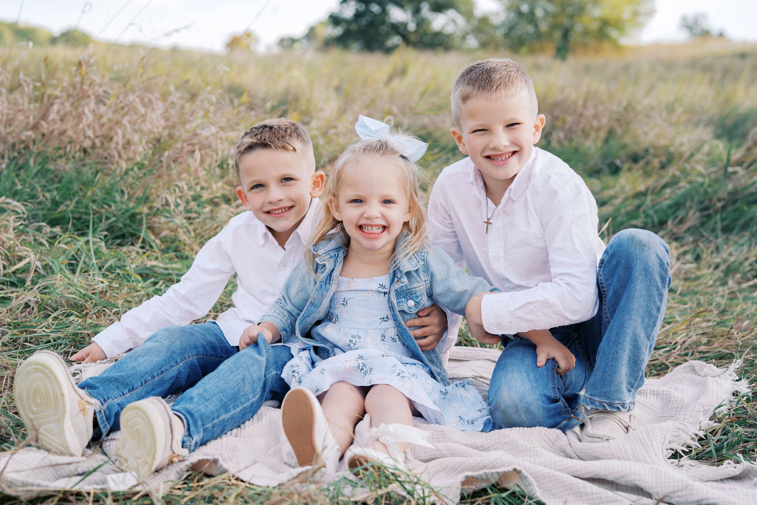 Three children sit close together on a blanket in a field wearing coordinated white and denim outfits.