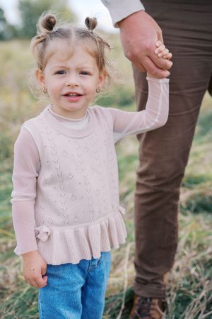A small child in a white sweater and jeans holds hands with someone while walking outdoors.