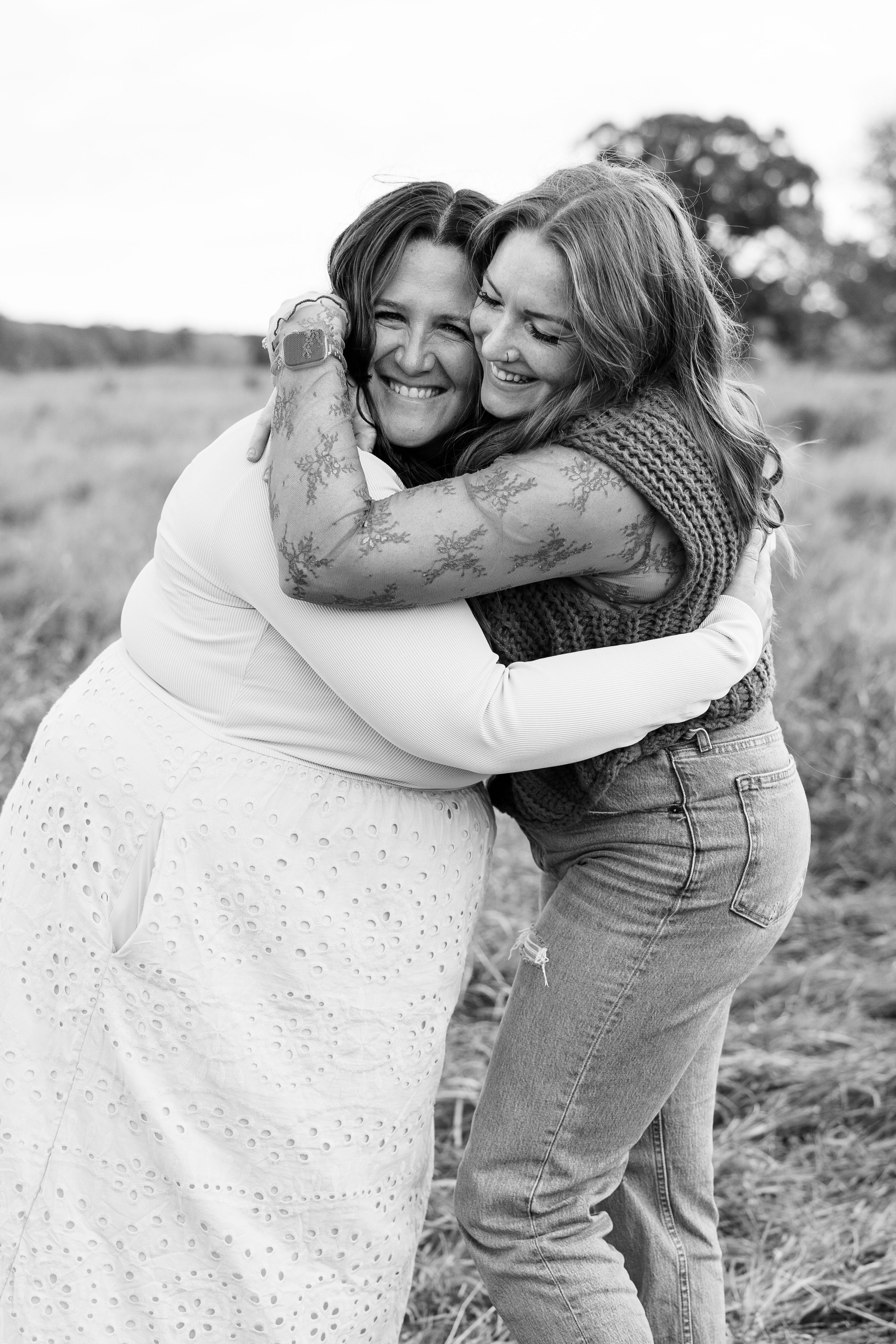 Two women share a warm embrace while laughing together in a black and white portrait.