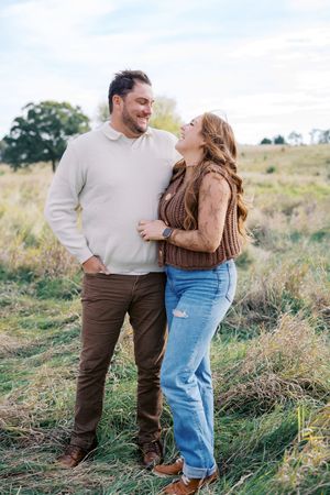 A couple embraces while standing in a grassy field on a sunny day wearing casual fall attire.