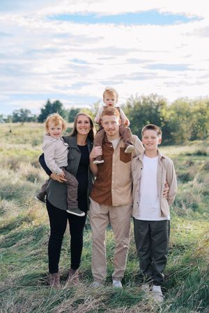 A family poses together in a grassy field on a sunny day wearing coordinated neutral-toned outfits.