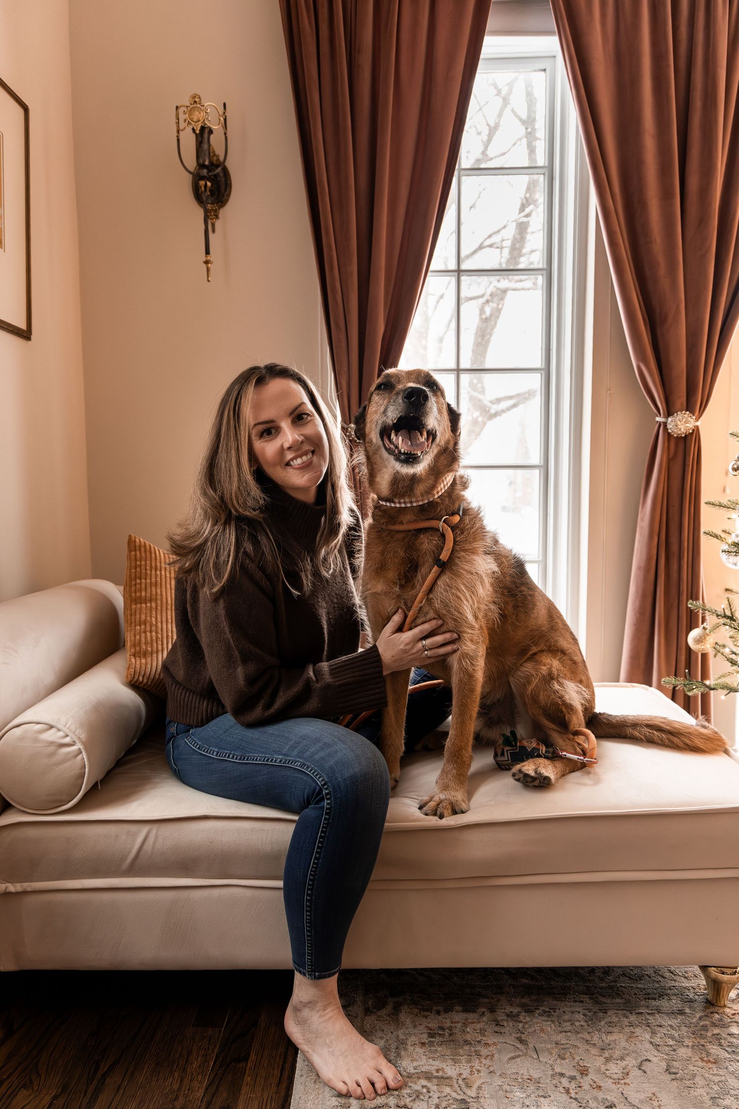 A person sits with their dog on a window seat in a room with brown curtains and natural lighting.
