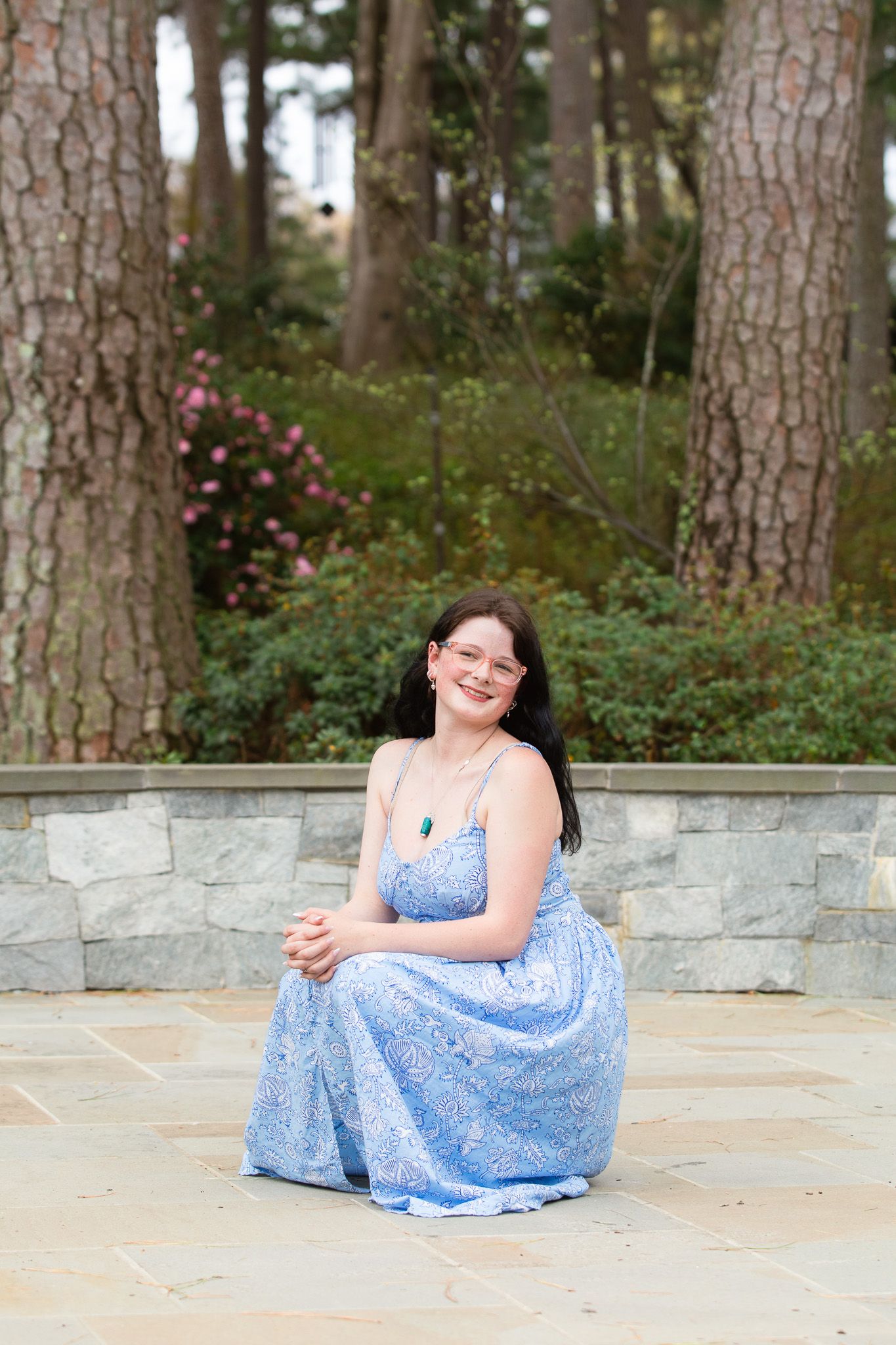 Young woman in blue floral maxi dress sitting on stone patio surrounded by lush garden trees and pink flowers at WRAL Azalea Garden