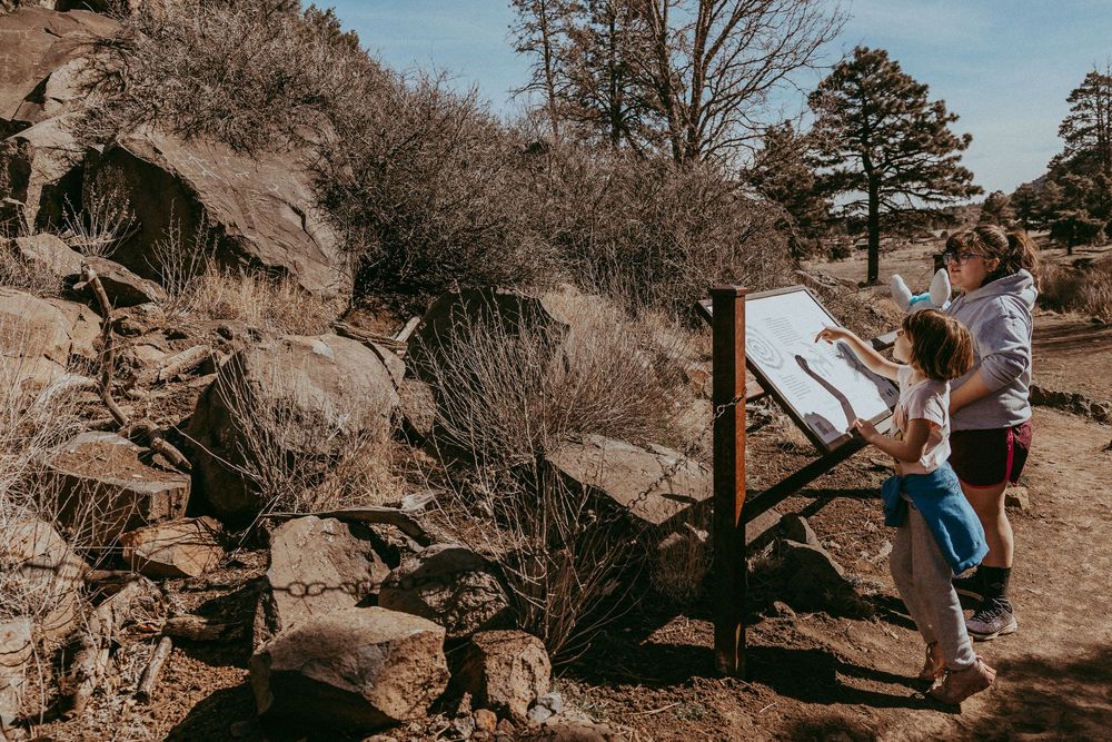 Two people studying a trail map on a wooden sign board in a rocky mountain landscape with pine trees. Whitney Popp, Popp Photography