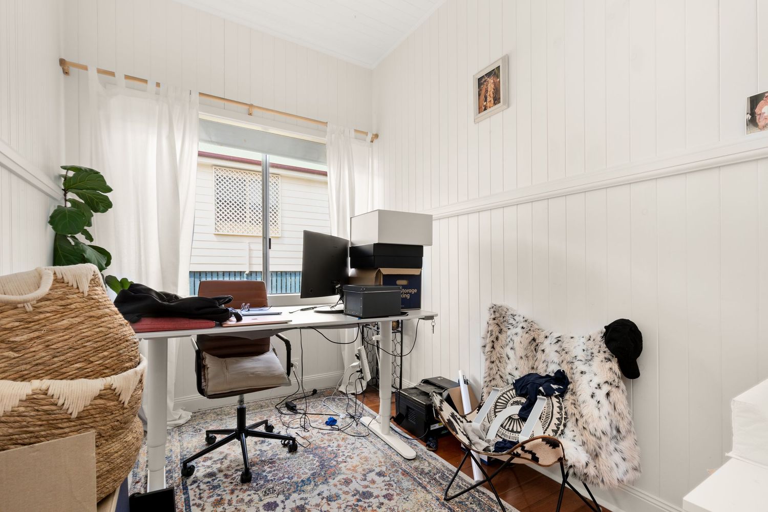 Small home office with standing desk, dual monitors, patterned rug, and white panelled walls.