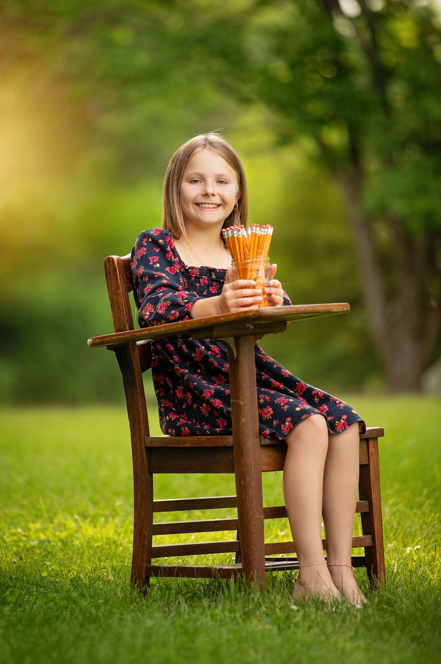 A child in a floral dress sits in a wooden chair on green grass holding a glass of pencils on a sunny day.
