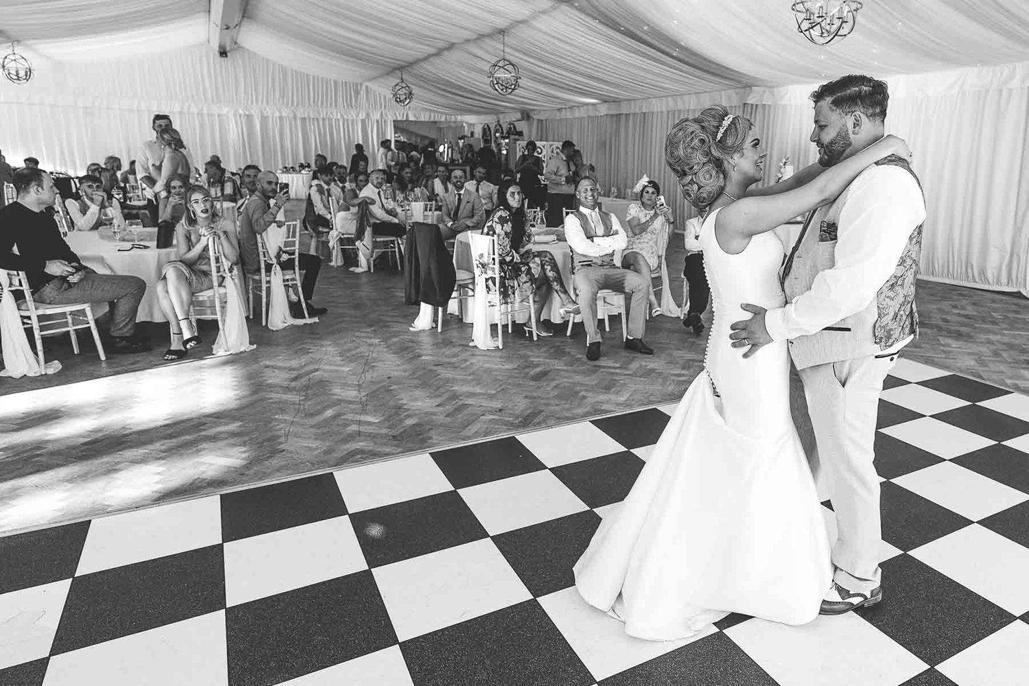 Bride and groom share first dance on checkered floor in elegant marquee wedding reception.