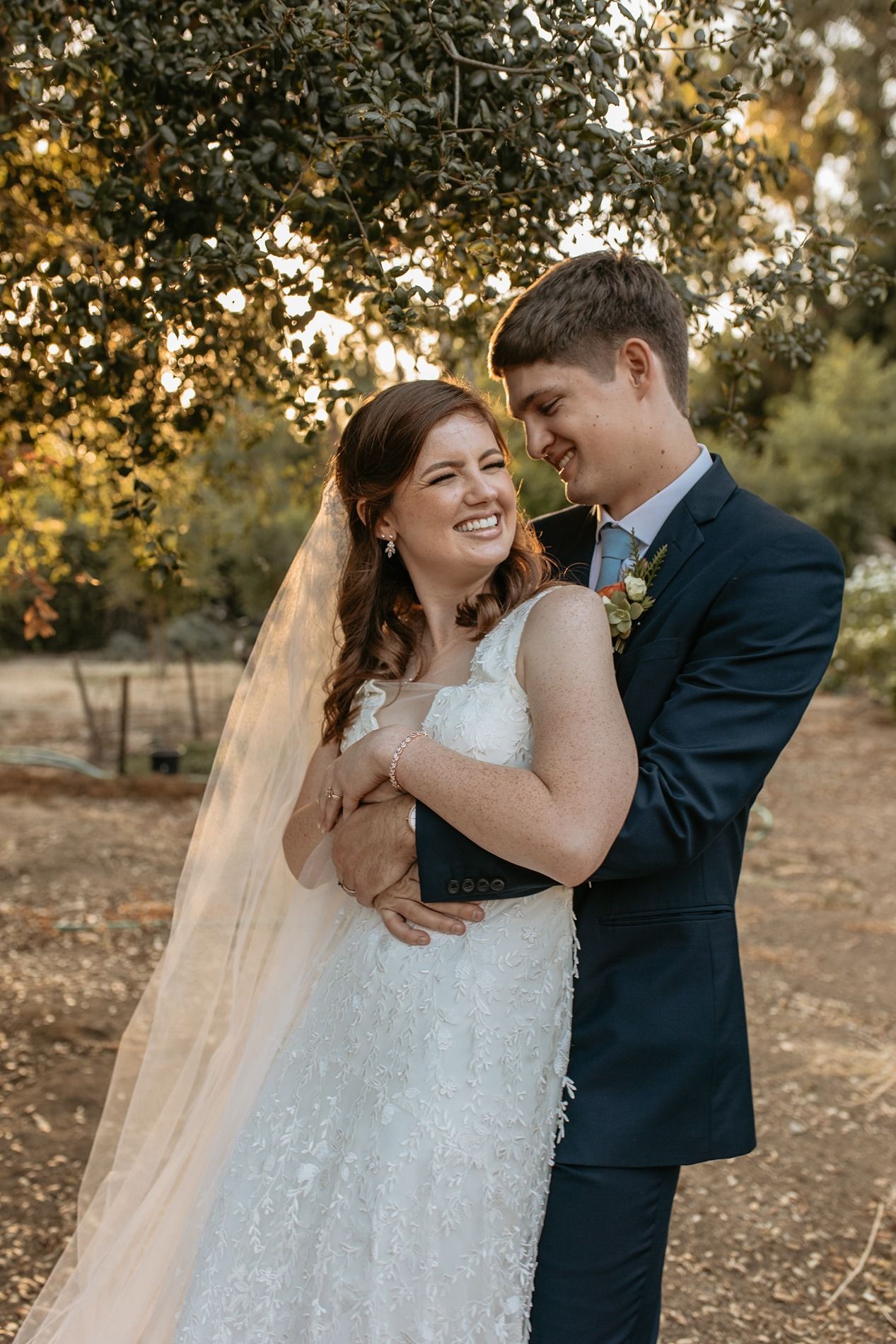 A smiling bride in a lace gown and veil embraces her groom in a navy suit outdoors at golden hour at Rancho Buena Vista Adobe.