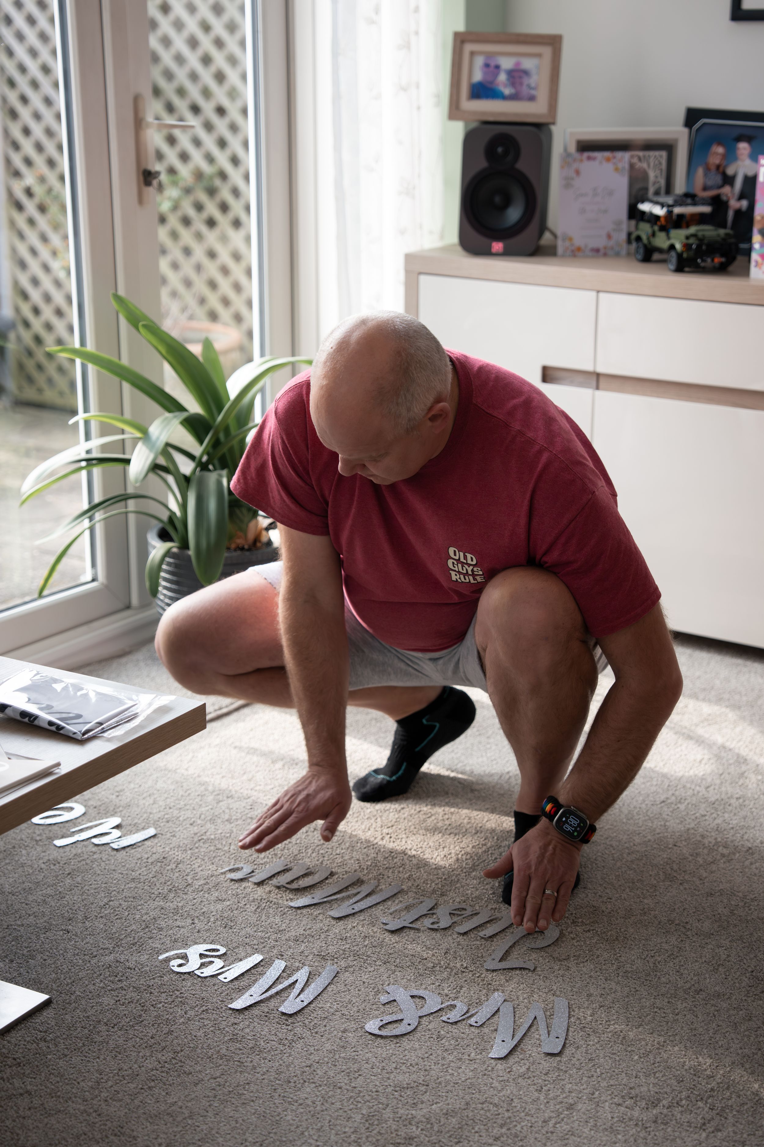Person in a red shirt crouches on carpet arranging letters or text pieces on the floor near a houseplant and window.