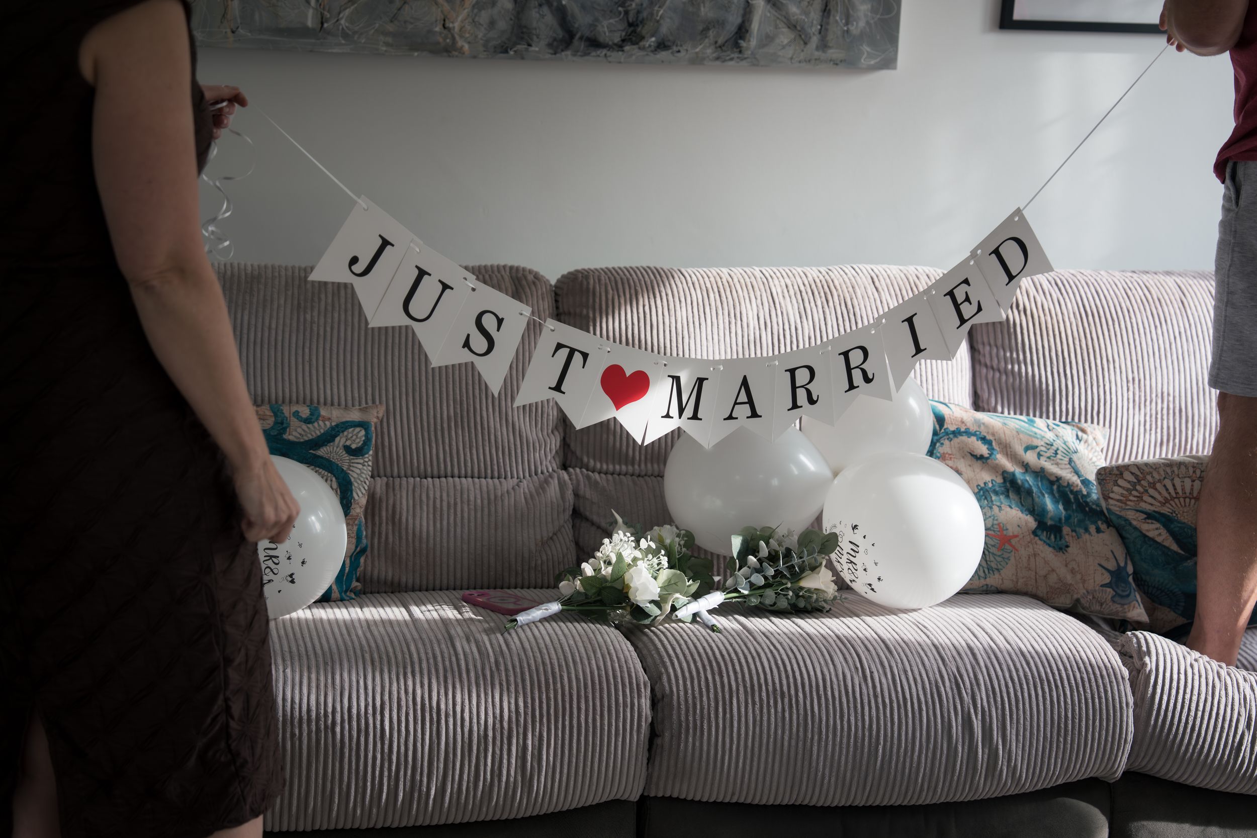 A 'Just Married' banner with a heart hangs above a gray couch decorated with white balloons and greenery.