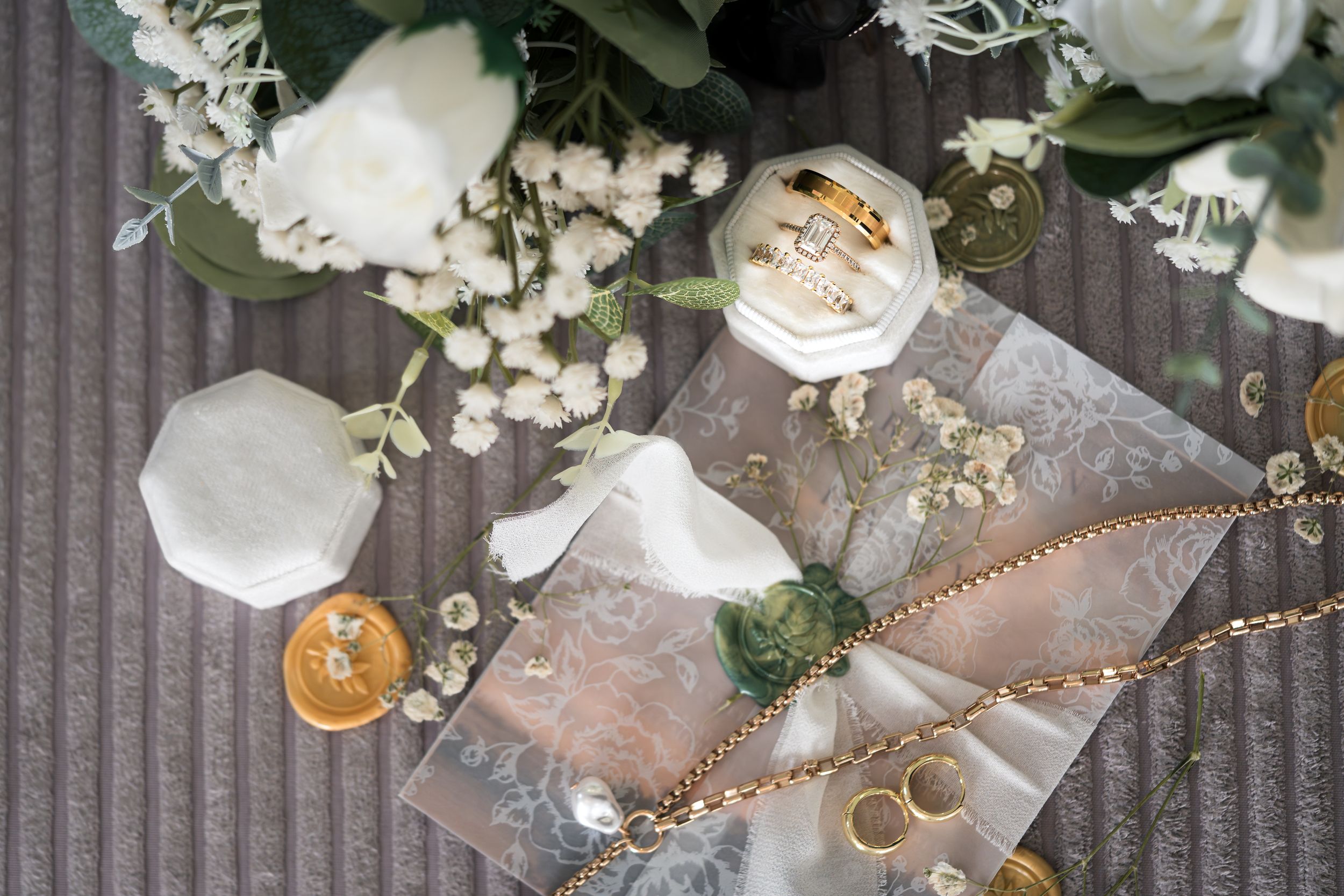 Wedding rings and floral arrangements with white roses and delicate greenery on a gray textured surface.