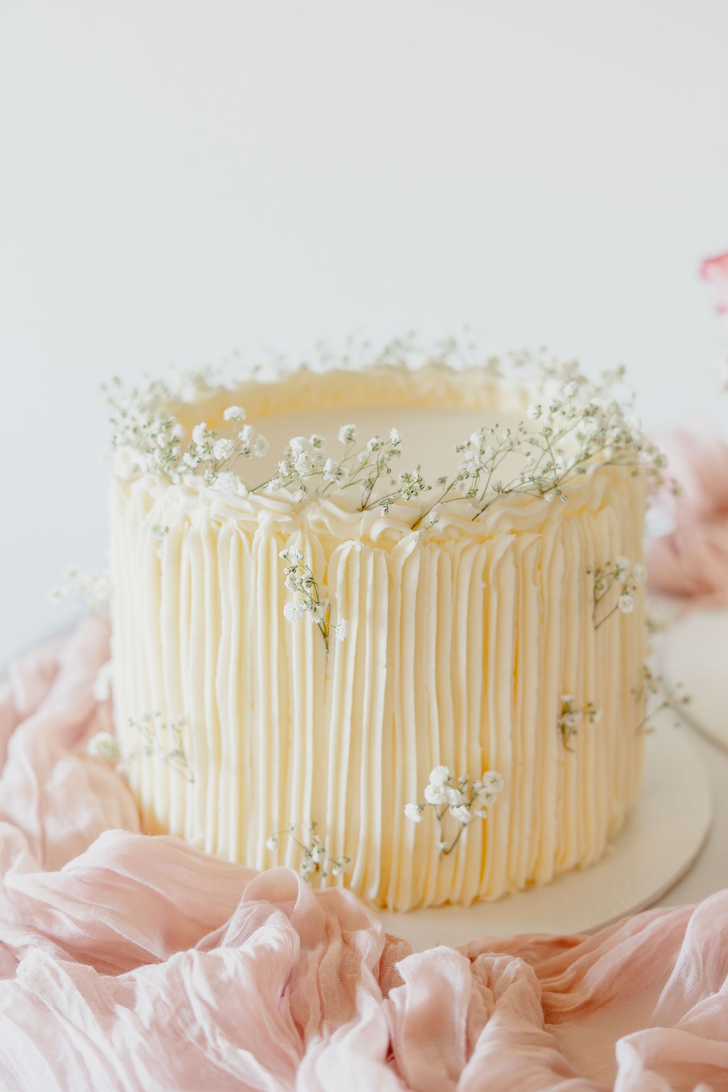 Elegant white buttercream cake with vertical ridges and delicate baby's breath flowers on a pink fabric backdrop.