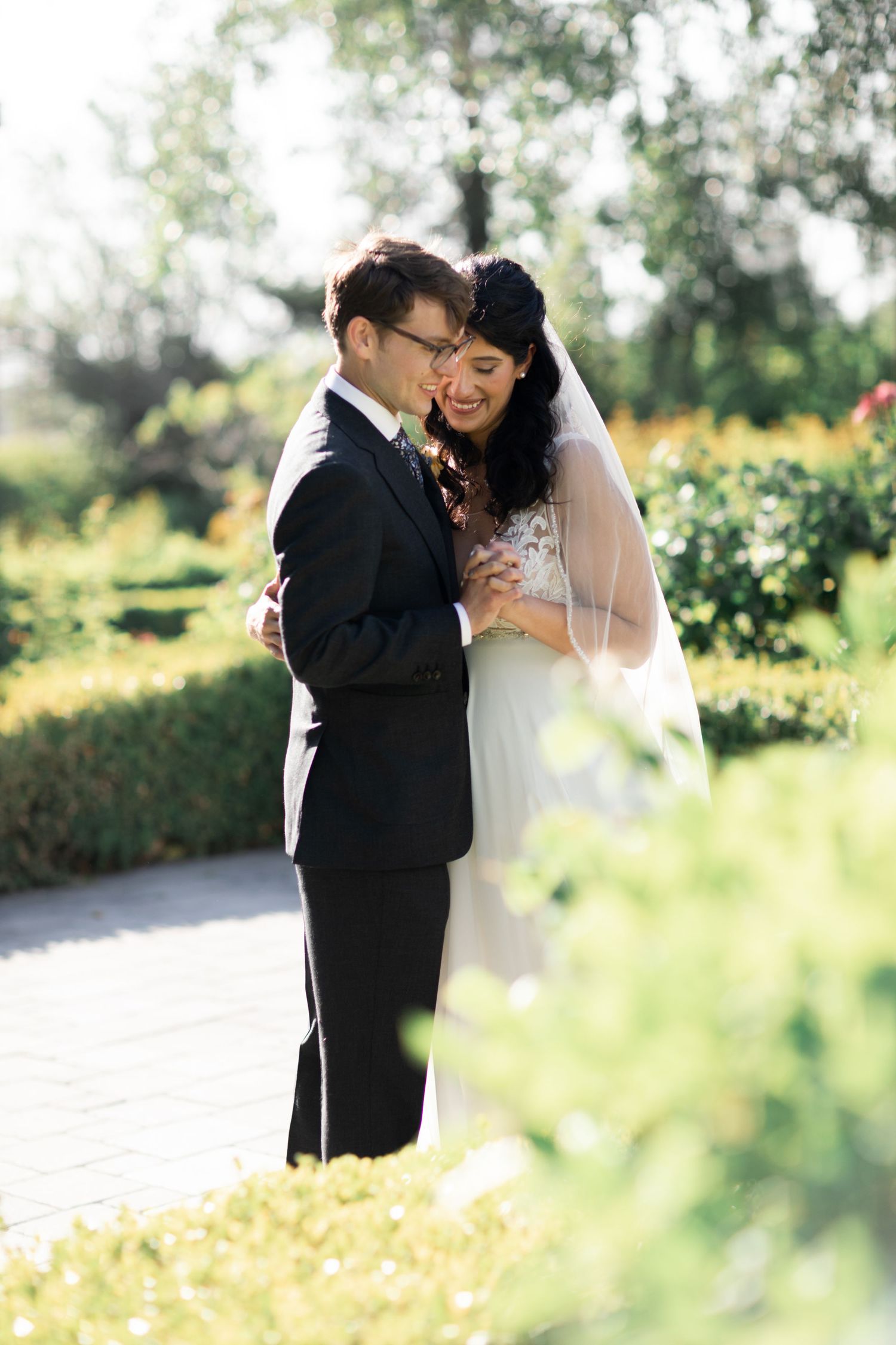 A bride and groom share an intimate moment in a sunlit garden with lush greenery and flowers in the background.