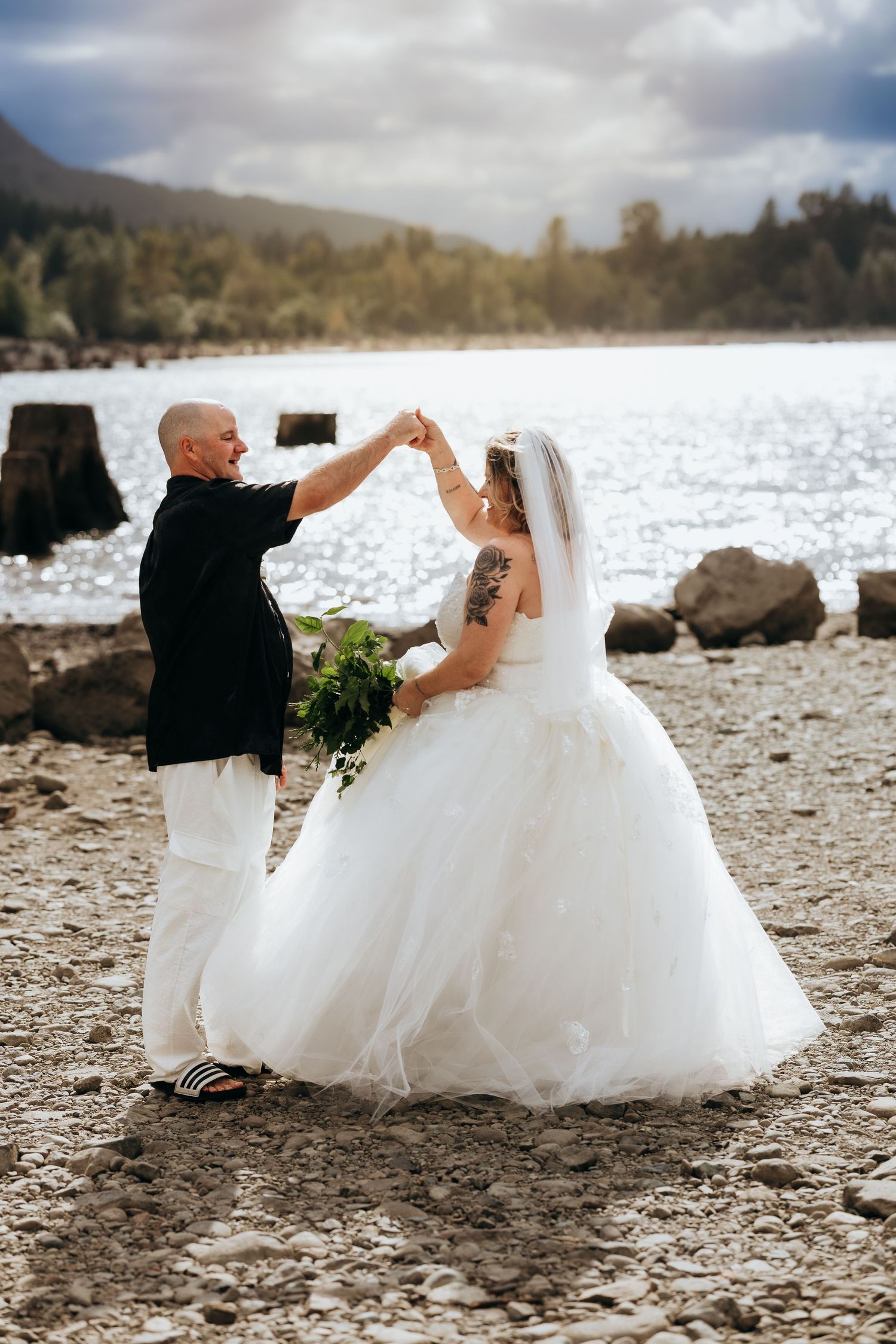 A couple in wedding attire dances together on a rocky beach shore with mountains and water in the background.