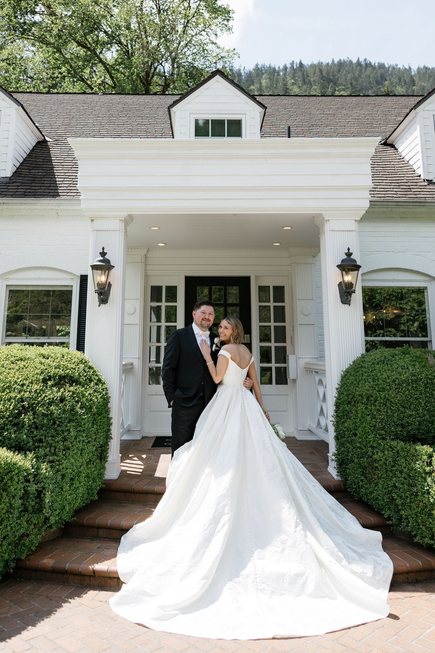 Bride and groom pose before a white colonial mansion entrance, her elegant gown trailing on brick steps.