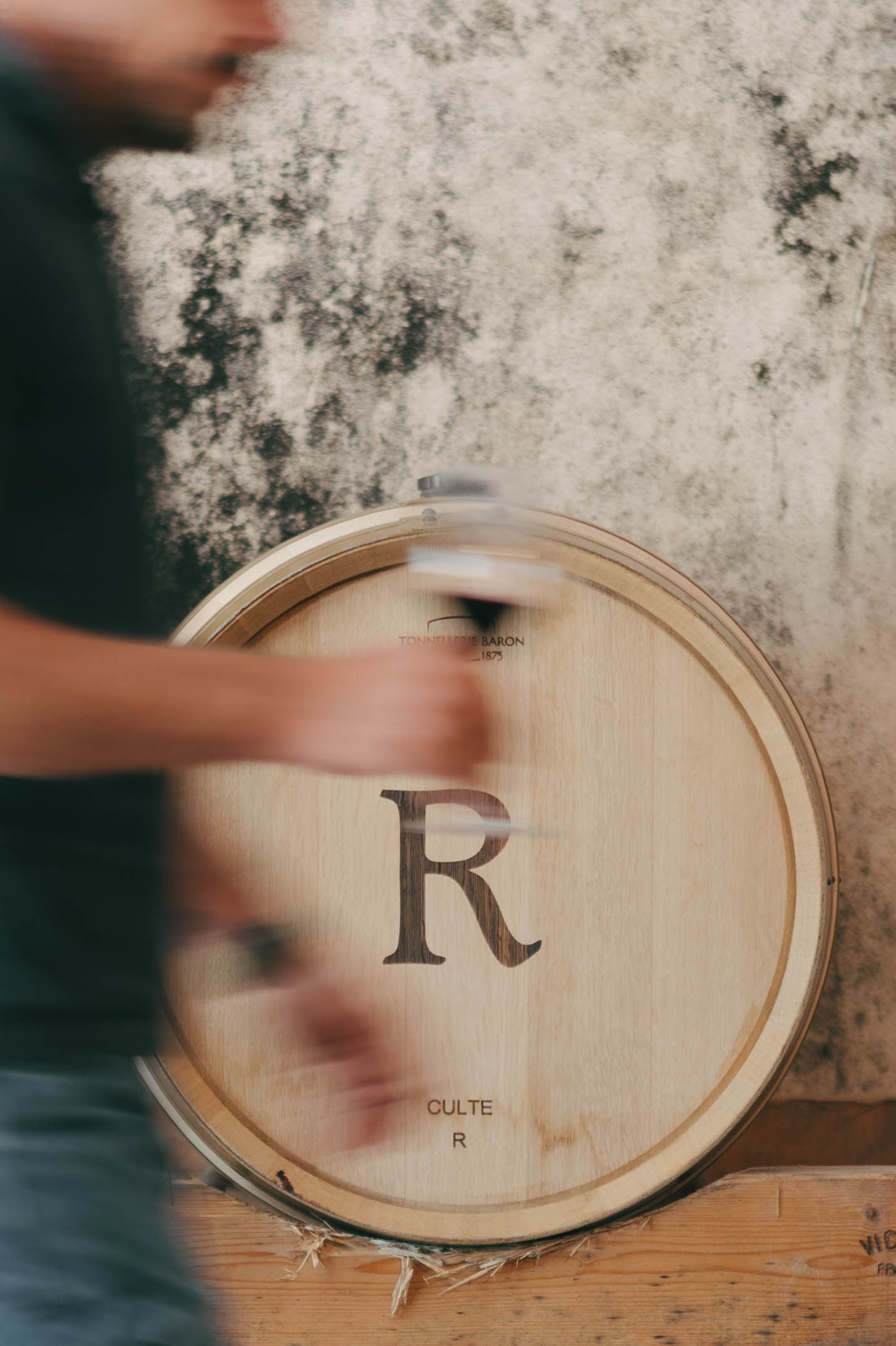 A wooden wine barrel engraved with letter R and text CULTE R, with a blurred person moving beside it.