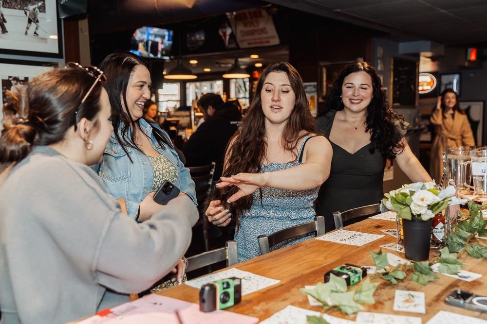 Women socializing and laughing at a bar counter with drinks and floral decorations during a casual indoor gathering.