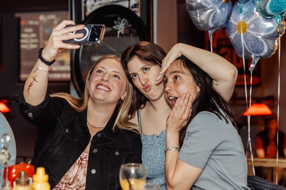 Three women pose for a fun selfie at a party, with colorful balloons in the background.