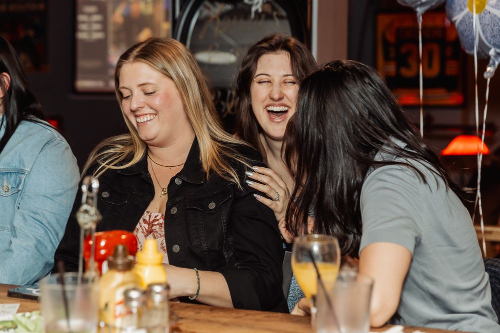 Three women laughing together at a restaurant table, enjoying drinks in a warm, lively bar setting.