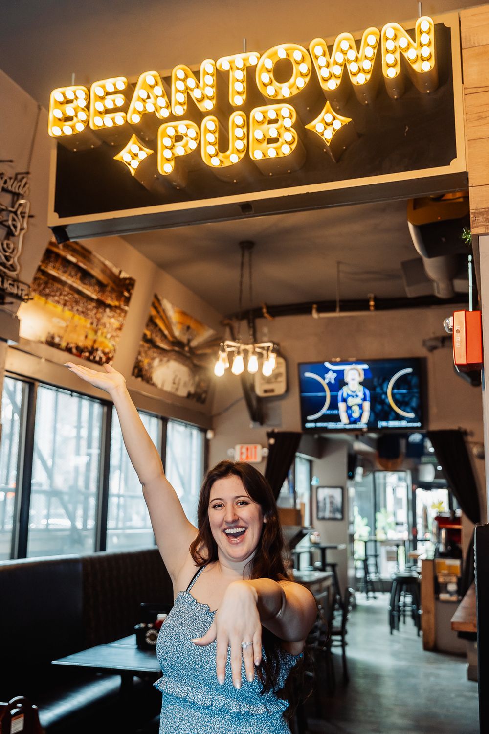 Excited woman posing under illuminated Beantown Pub sign in Boston bar, raising her arm in celebration.