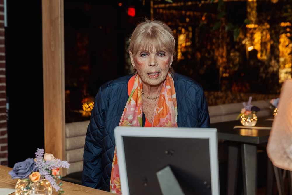 Older blonde woman in blue jacket and colorful scarf sitting at a desk with a monitor in a warmly lit venue.