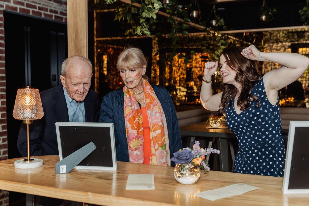 Elderly couple reviewing photos on tablets at a restaurant counter while a young woman celebrates nearby.