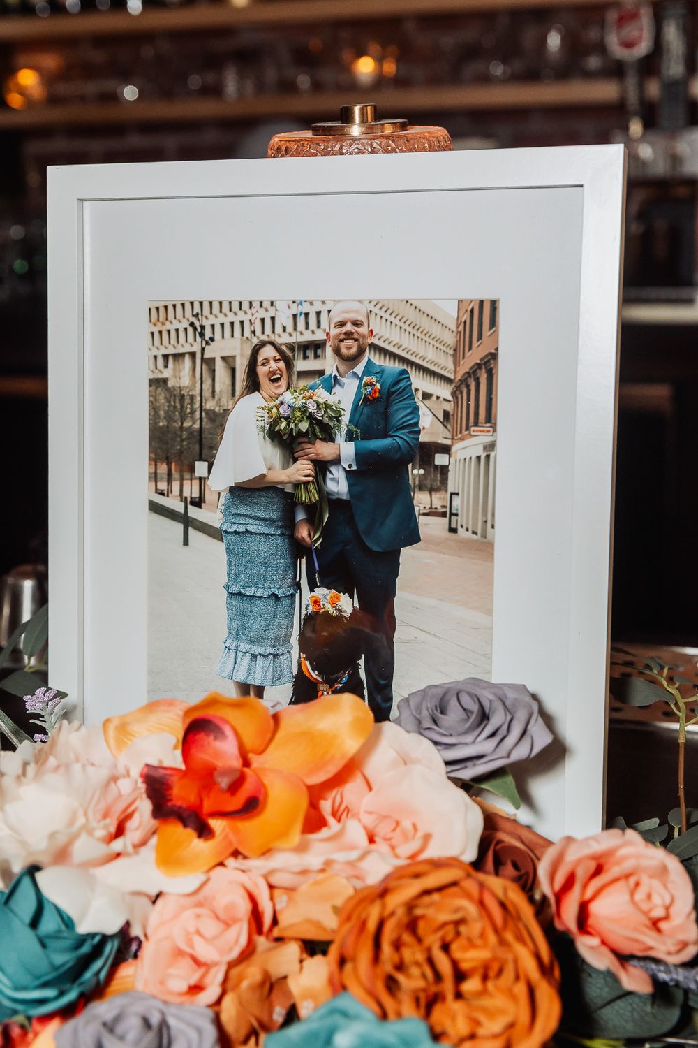 Framed wedding photo displayed with colorful floral arrangement featuring orange orchids and pink roses at a reception.